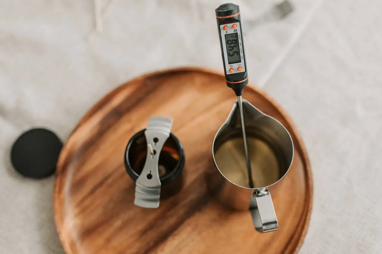 A flat lay of candle making tools including a jar, wick, and digital thermometer.